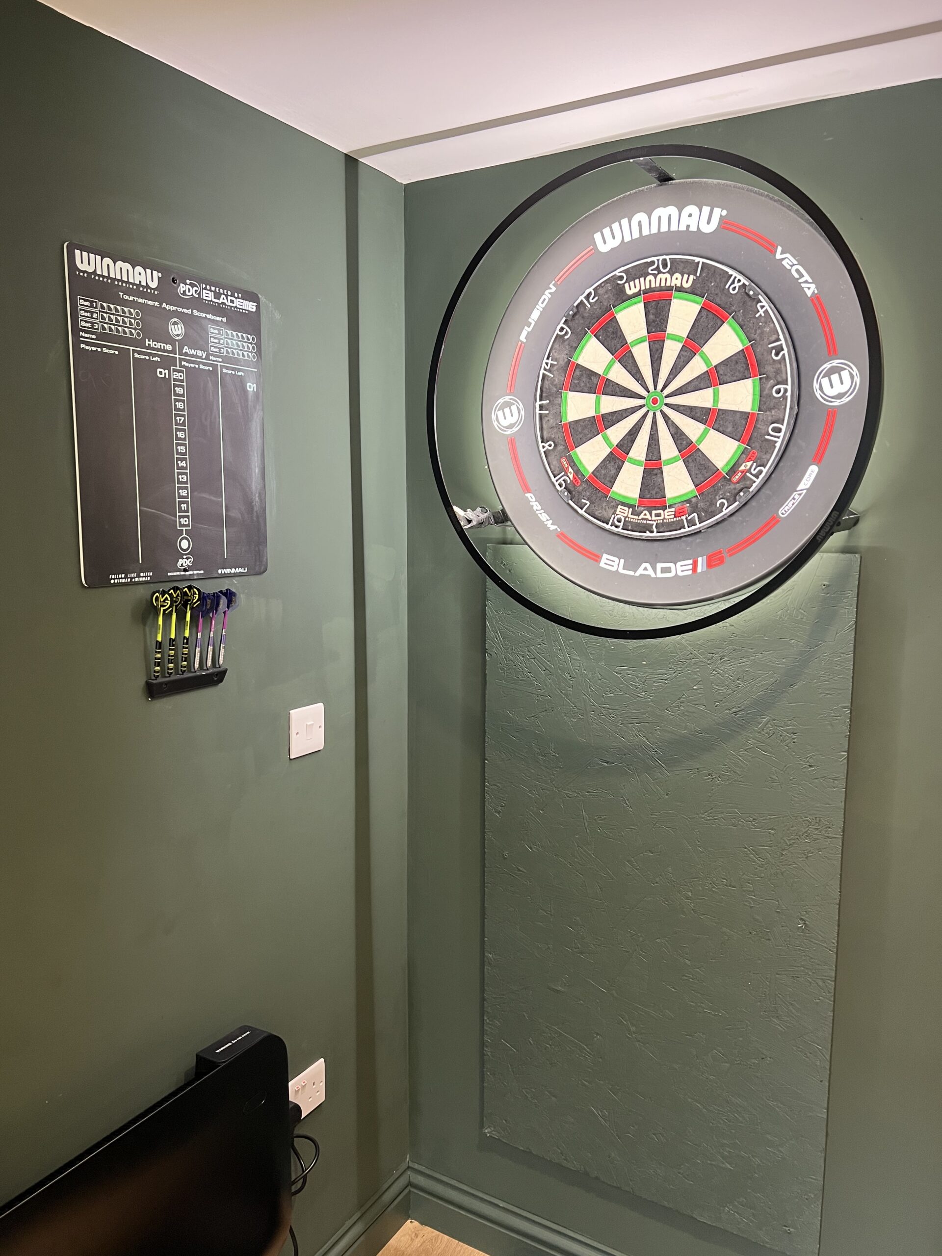 Interior wall of garden bar room with Winmau dartboard, LED surround and scoreboard on dark green painted wall