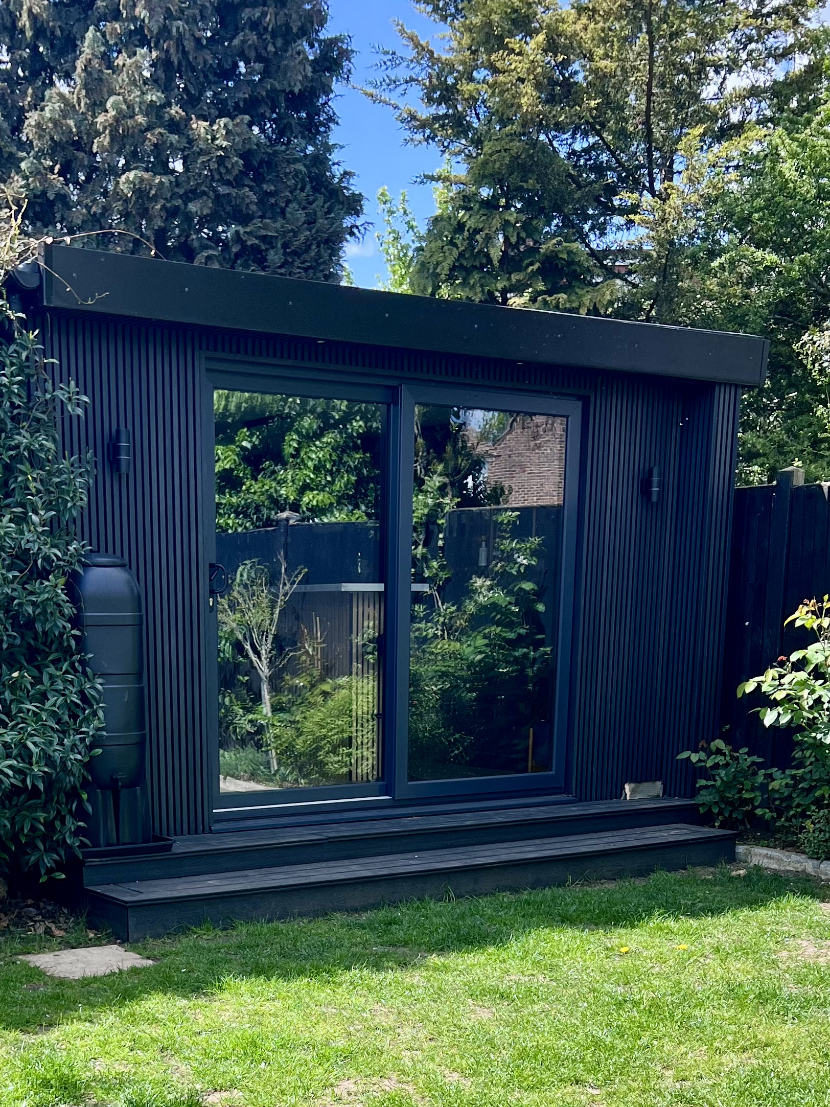 Garden bar room exterior in daylight, black composite cladding and full-width sliding aluminium doors, Surrey garden