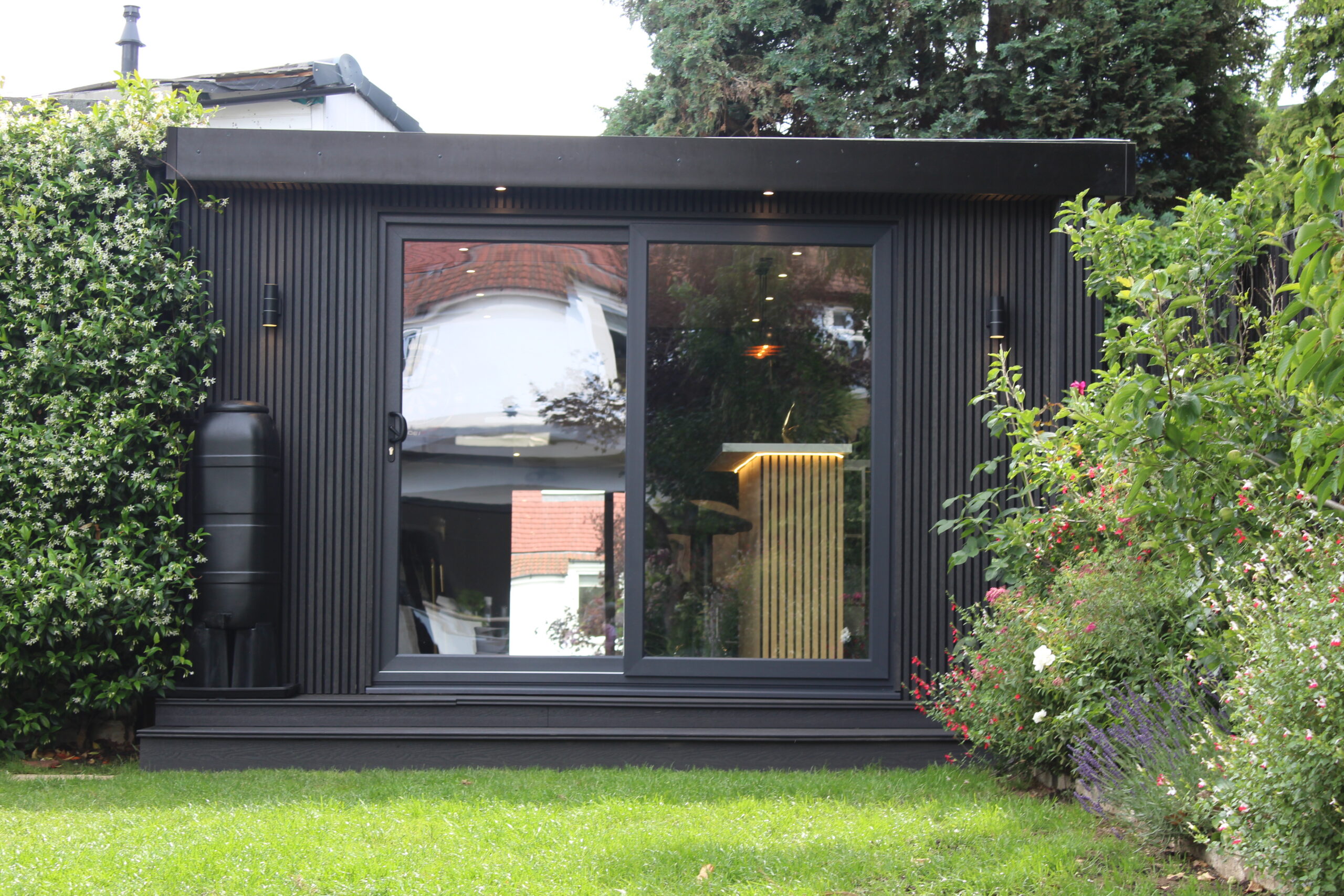 Anthracite slatted-cladding garden room with full-width sliding glass doors and interior lighting, set among jasmine, roses and lavender — My Retreat Garden Rooms, Surrey