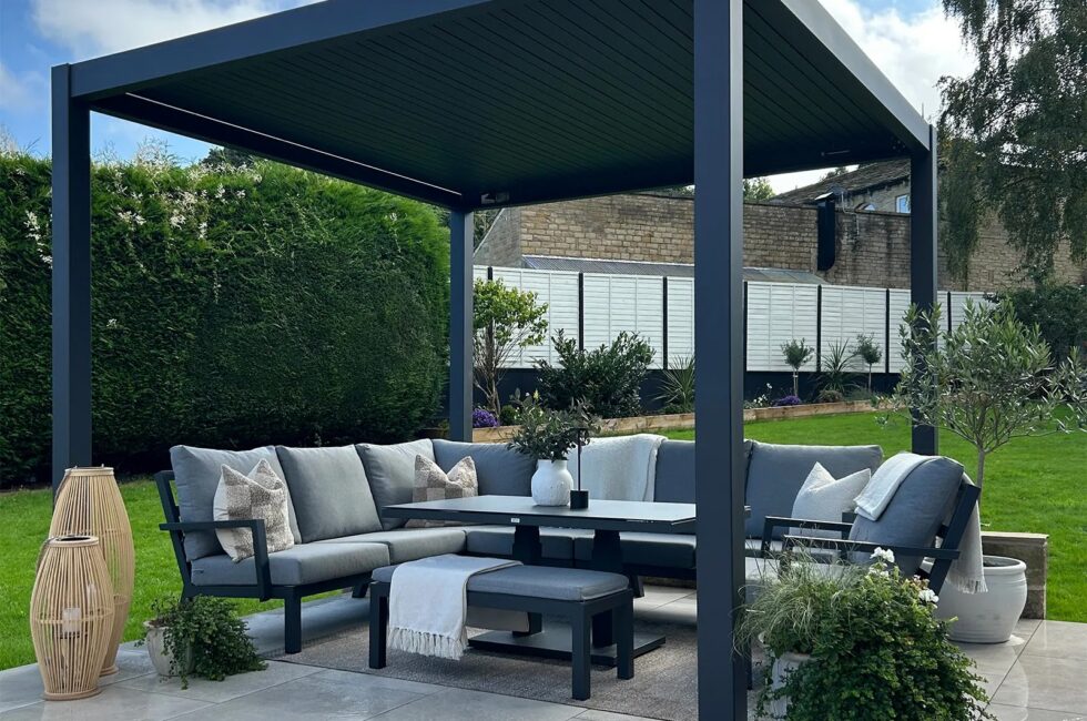 Grey aluminium pergola with a louvred roof over a patio seating area with a corner sofa and dining table